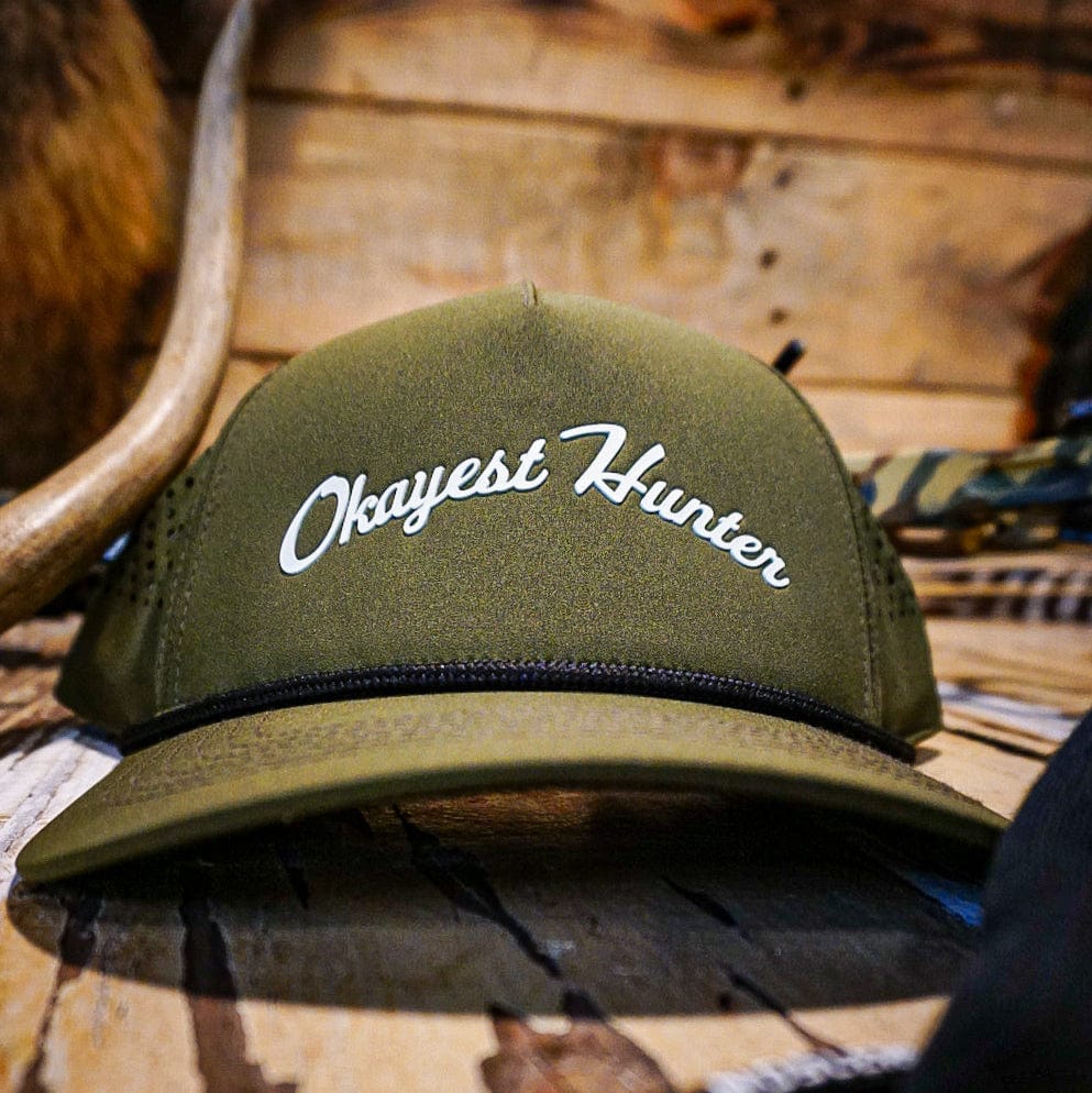 Green cap with 'Okayest Hunter' text on a wooden surface with hunting equipment.