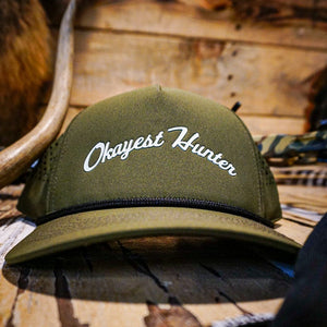 Green cap with 'Okayest Hunter' text on a wooden surface with hunting equipment.